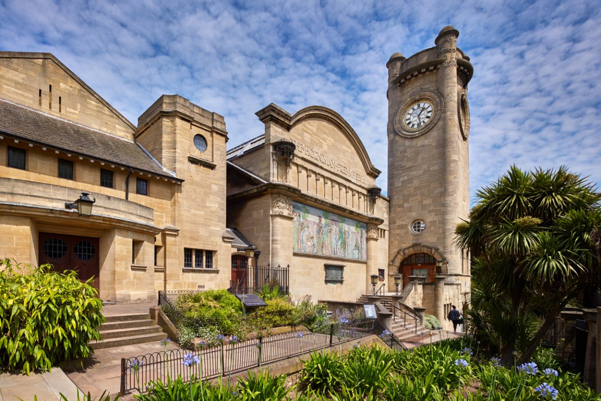 A shot of the exterior of the Horniman - a large pale yellow stone clocktower with rounded edges, a building next to it with a curved roof and a mosaic, and a third structure closer to the camera with a peaked roof, made of similar stone. There is a path along the front of all three with flower beds featuring green and purple flowers in front of the camera. The sky is blue