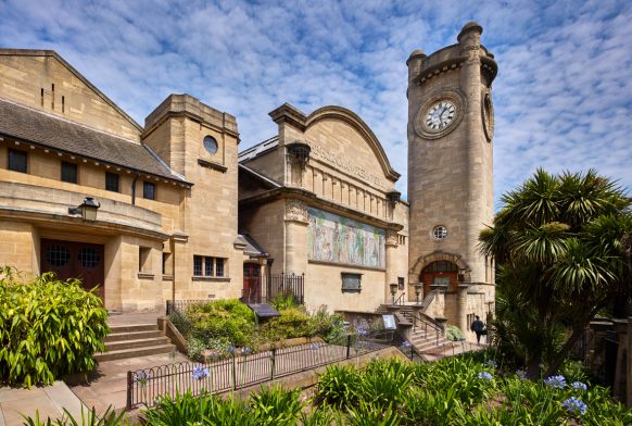 A shot of the exterior of the Horniman - a large pale yellow stone clocktower with rounded edges, a building next to it with a curved roof and a mosaic, and a third structure closer to the camera with a peaked roof, made of similar stone. There is a path along the front of all three with flower beds featuring green and purple flowers in front of the camera. The sky is blue