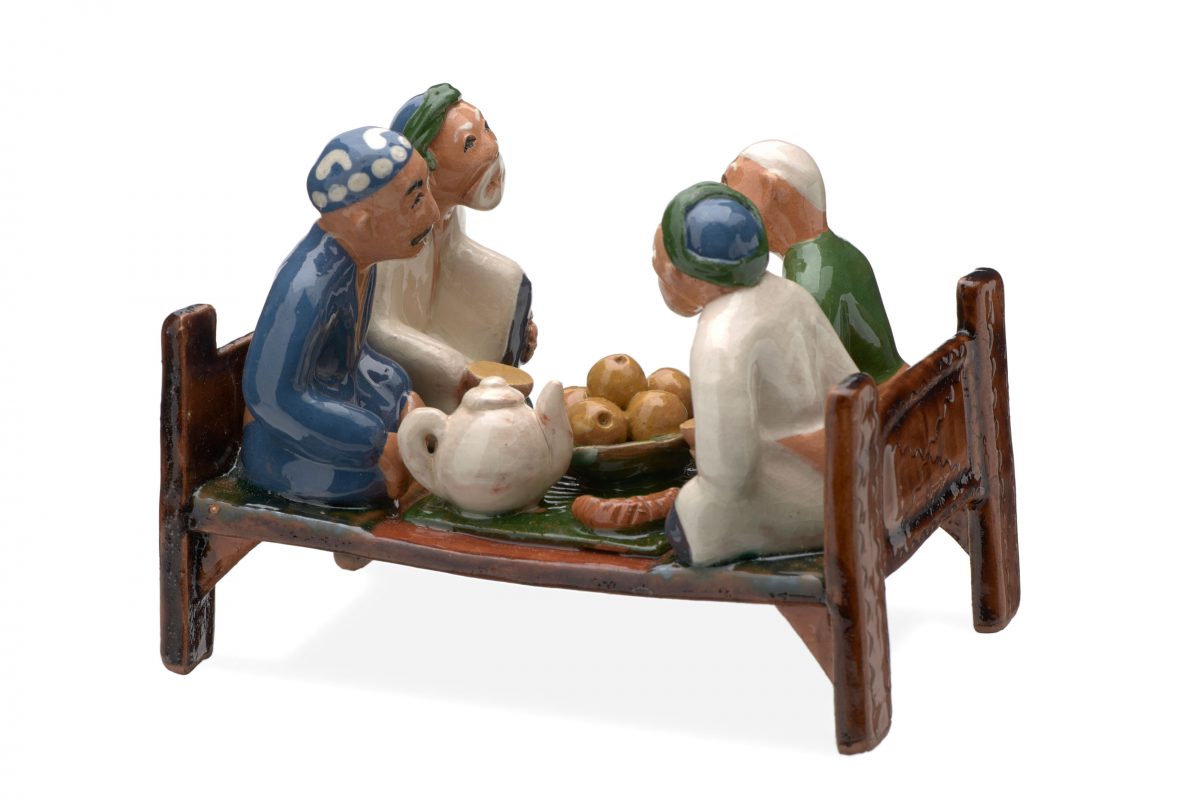 Ceramic model of four men, three ‘grey beards’ and one younger man, eating a meal, seated or kneeling on a bed-table, local name takhta around a green cloth with a bowl of apples, flat bread and a white teapot.