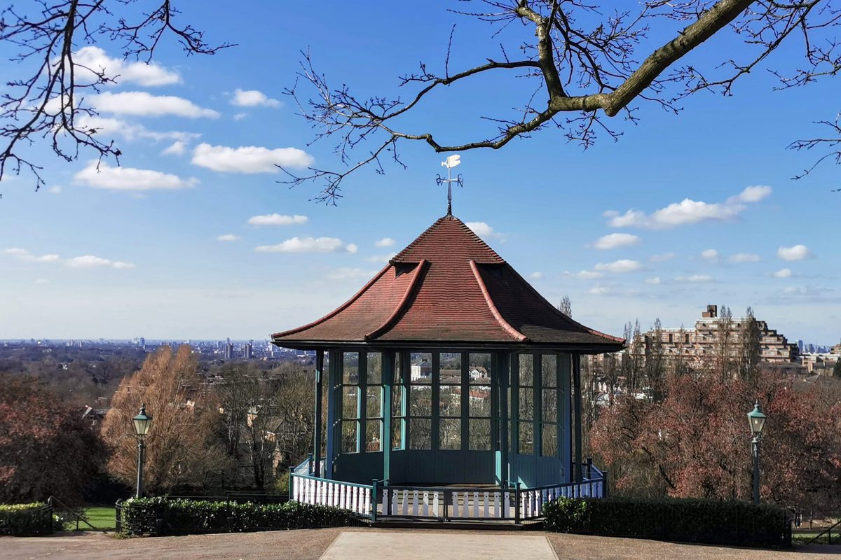 Small glass building stands in the middle of path. In the background the city can be seen and