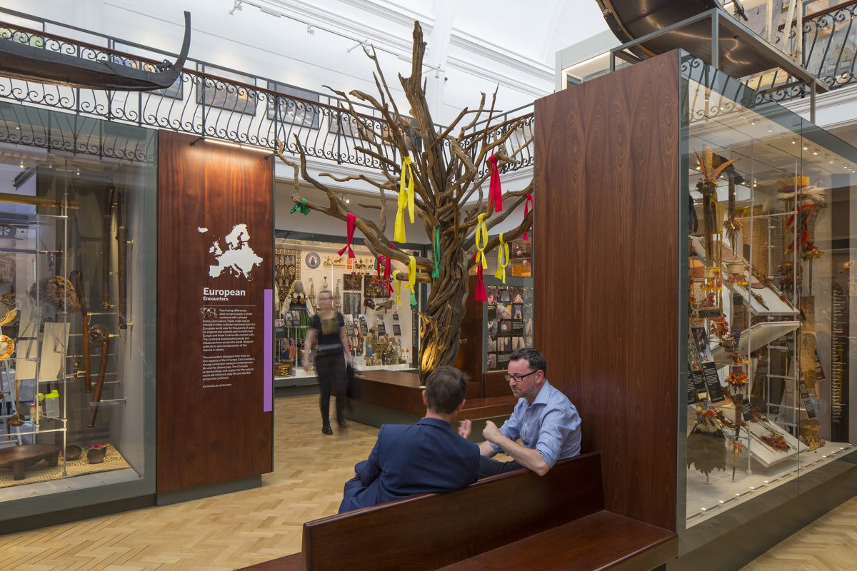 A tree with strips of fabric hanging from the branches sits in a gallery. Two men sit on a bench in the foreground and a woman is walking past the tree looking into display cases.