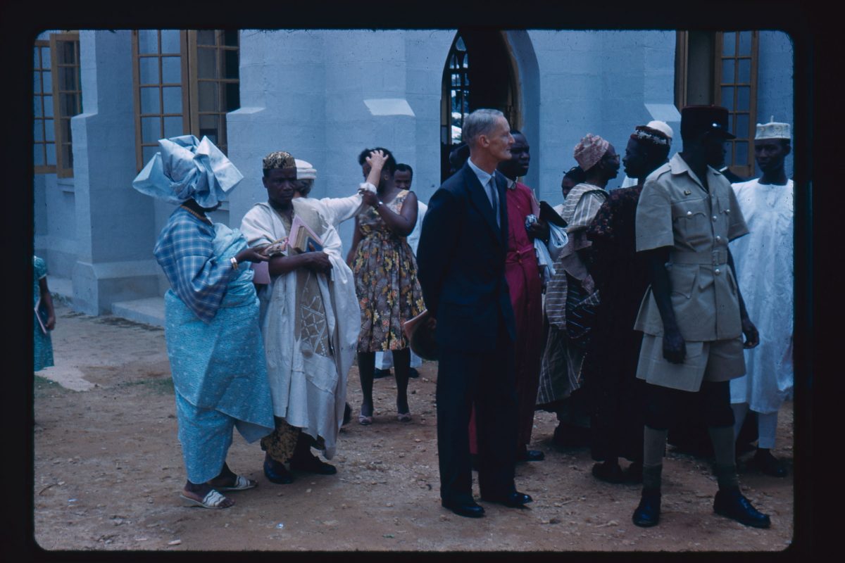 A old colour photo of a formal or celebratory occasion. Two people to the left are in blue and white clothes. A man to the centre is in a suit and to his right is a man in a khaki uniform. Behind him are a group of people wearing robes. Behind the people in the foreground is a woman in a sun dress who is holding a second woman's arm, who is in a white dress. The woman in the white dress is reaching for her and looking away from her at the same time