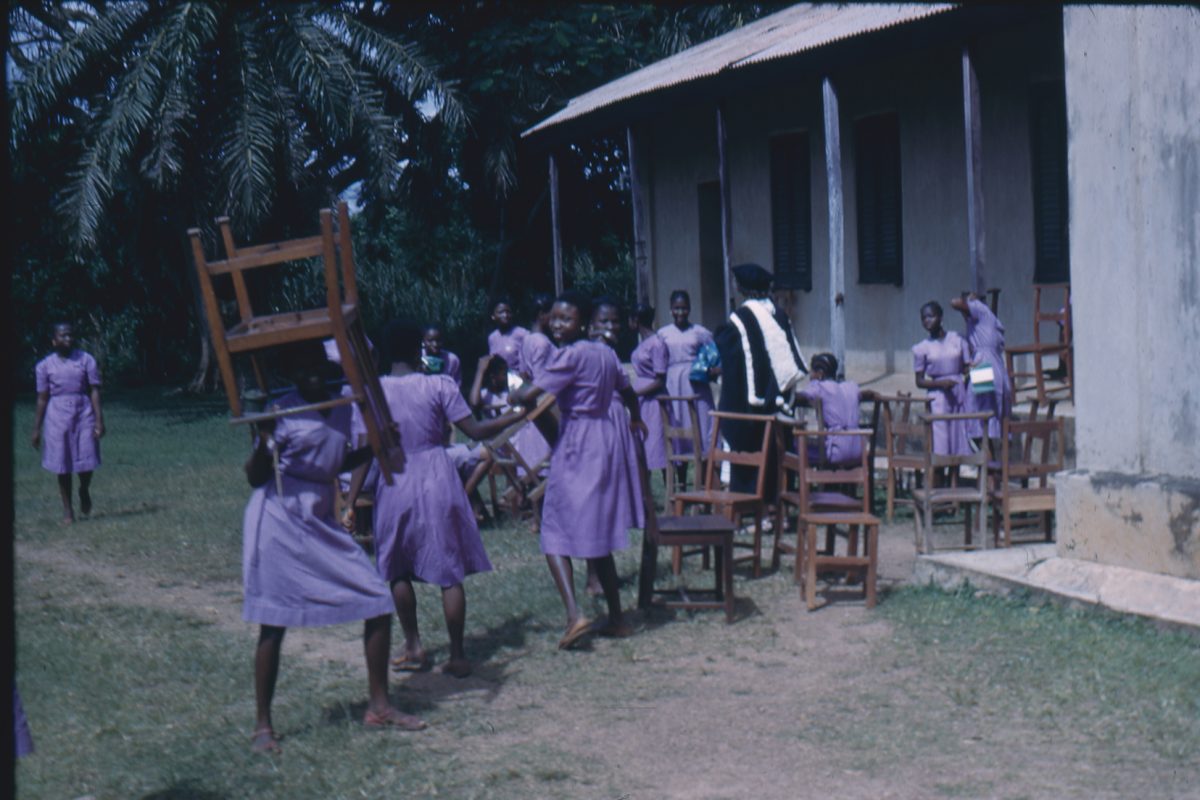 A group of school children moving chairs outside school building.