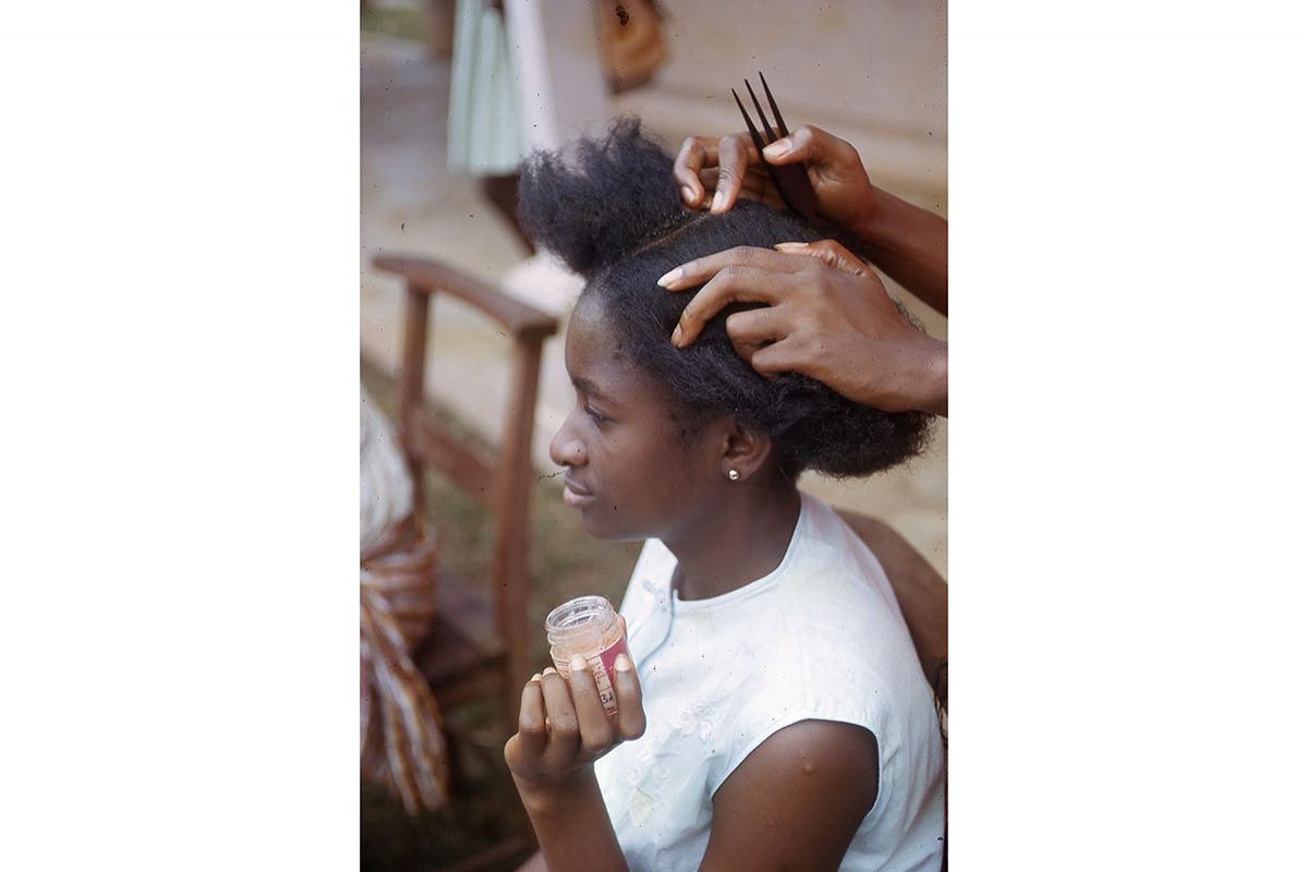 A woman is having her hair done - it has been parted by some hands (person unseen) who is also holding an afro comb. The girl wears a white top and is holding up a pot of hair product for the person doing her hair to use.