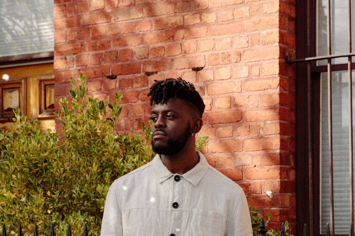 A man wearing a white shirt looks to the left against a backdrop of a red brick wall