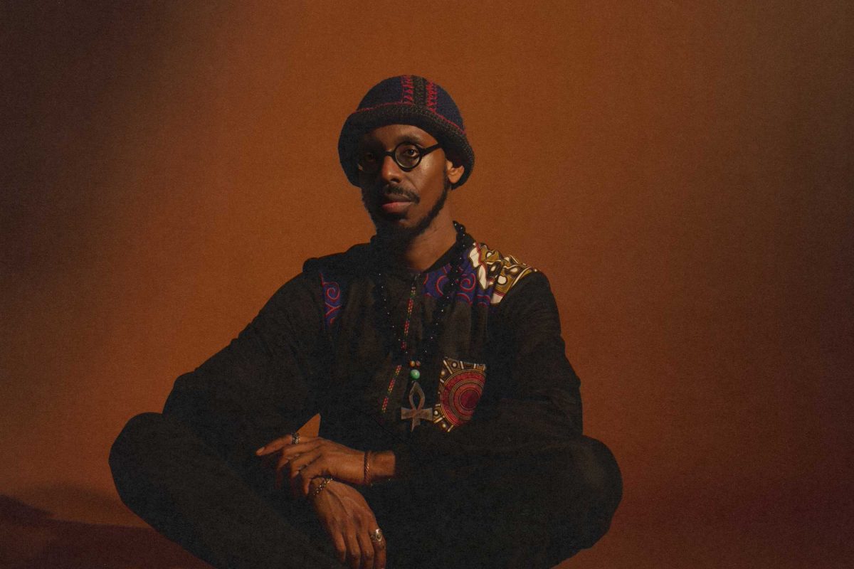 A man wearing glasses sits cross legged on the floor against a moodily lit red wall