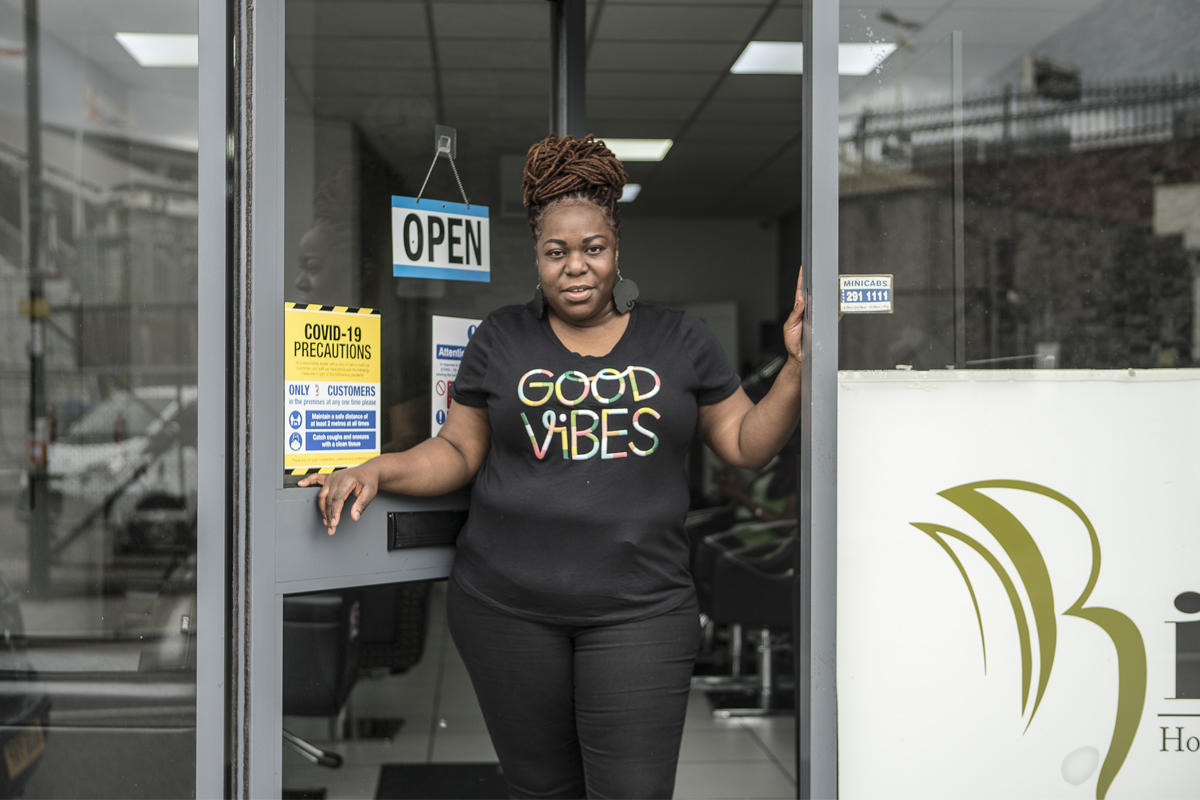 A woman wearing a t shirt that reads 'good vibes' stands in the door of a hair salon, looking at the camera