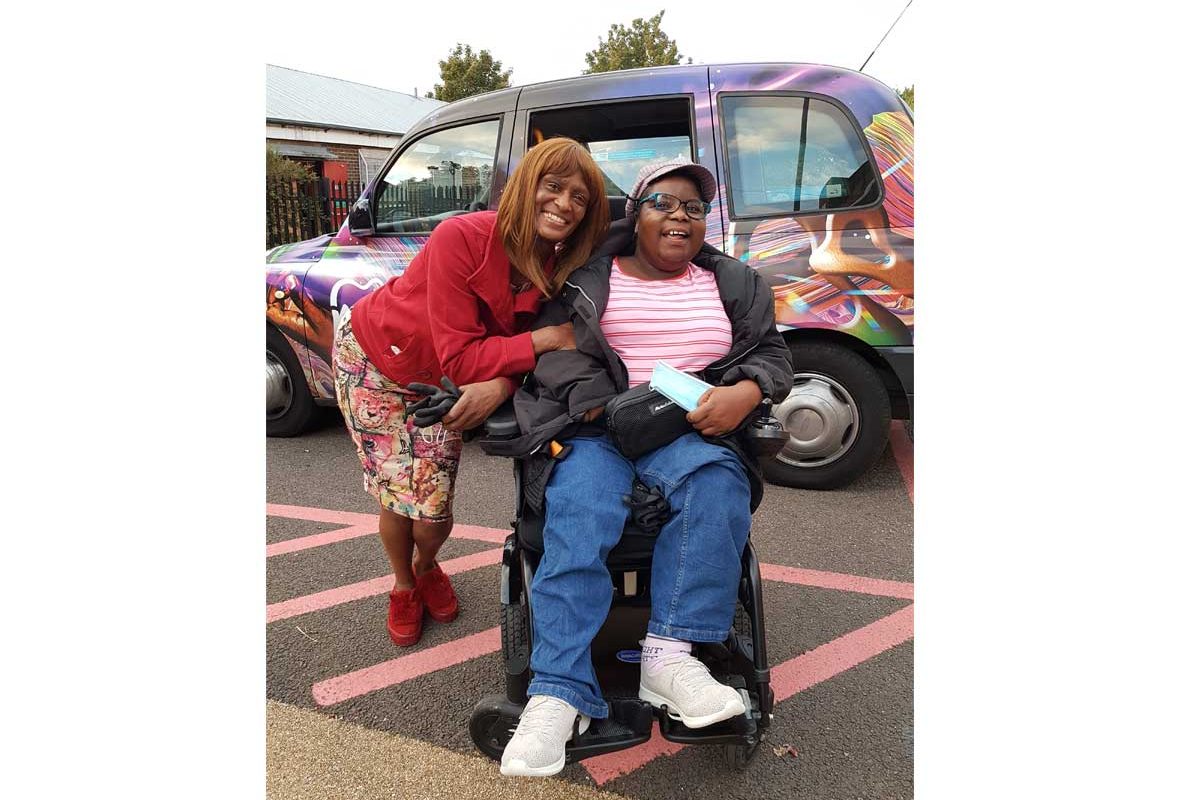 Tinu sits in a wheelchair and Heather stands next to her, both in front of a black cab