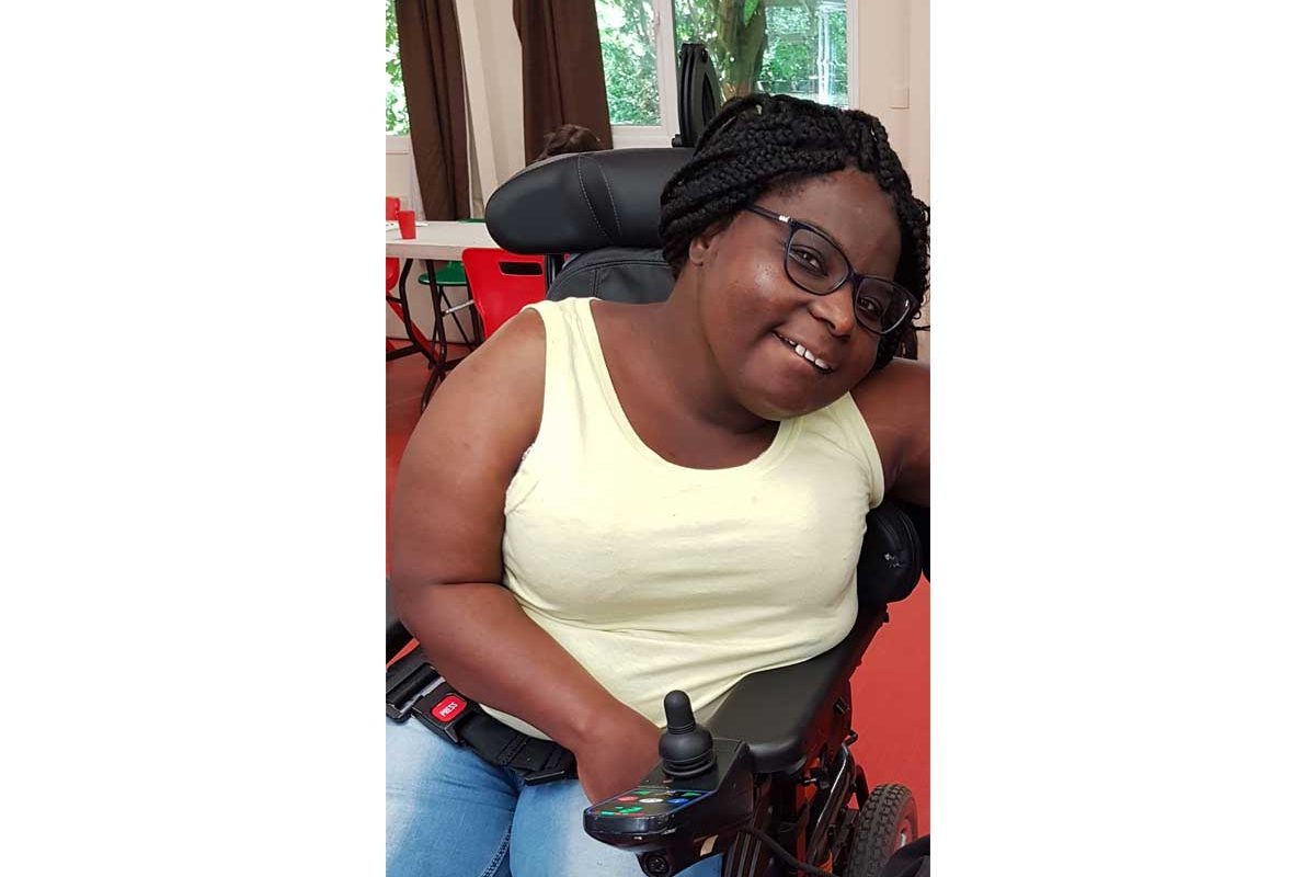 Tinu sits in a wheelchair and smiles at the camera. Her long hair is plaited back into a braid.