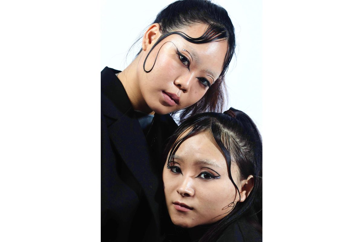 Two Asian women with long dark hair in ponytails stand with their heads close together. They have loose hair at the front of their heads, loosely gelled into swirls