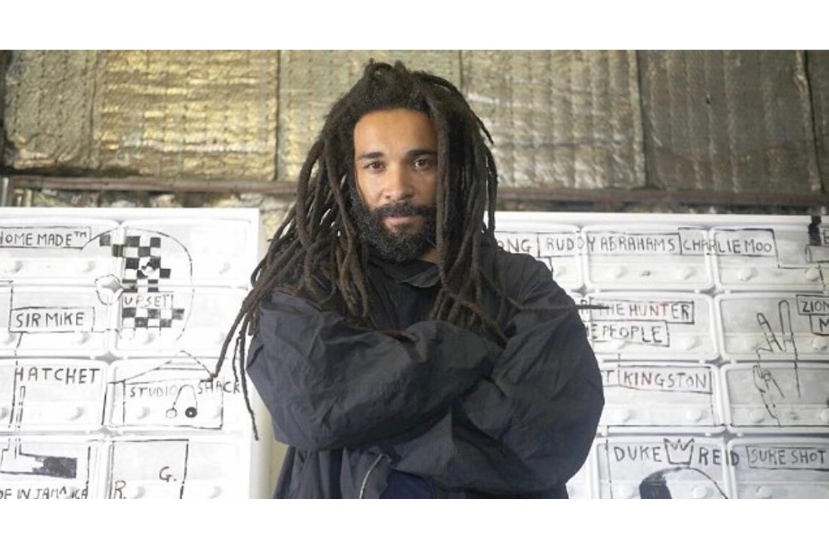 Andrew Ibi, a black man with dreadlocks looks at the camera with his arms folded. He is standing against a white wall with graffiti