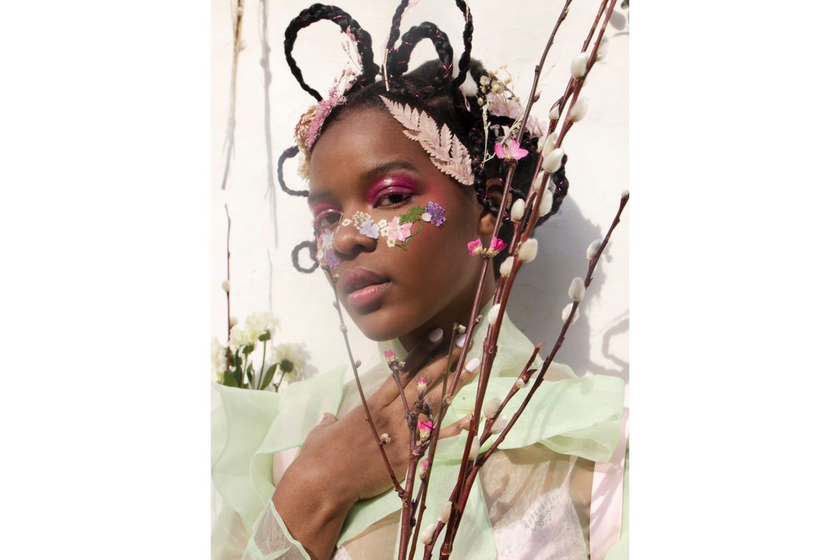 A Black woman wearing a pastel green floaty shirt and wearing flowers in her hair and on her face looks at the camera. She is surrounded by flowers