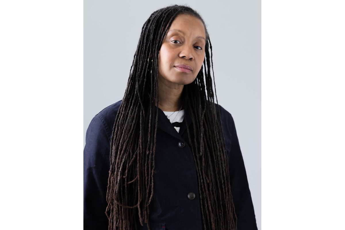 A Black woman, Sharon D Lloyd, looks at the camera. She wears a buttoned up cardigan and has long dreadlocks