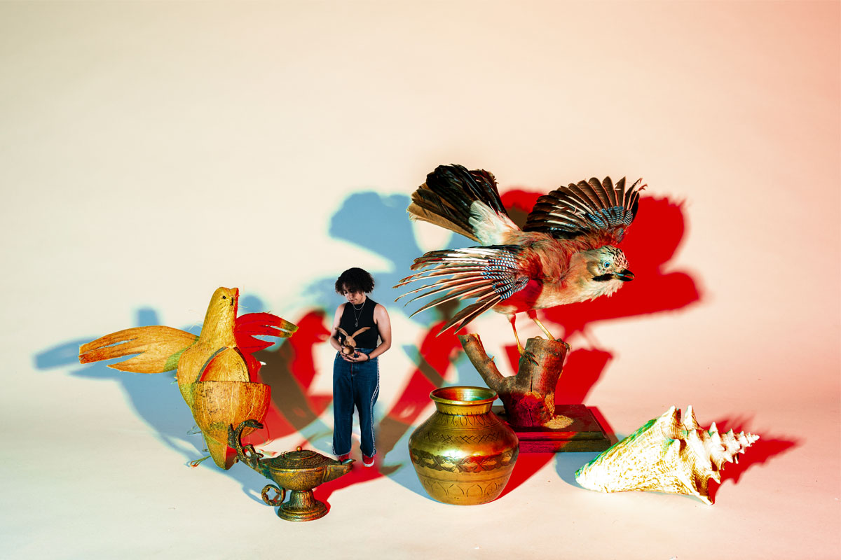 Woman wearing black top and jeans holds a bird model surrounded by giant models of birds, lamps and a shell