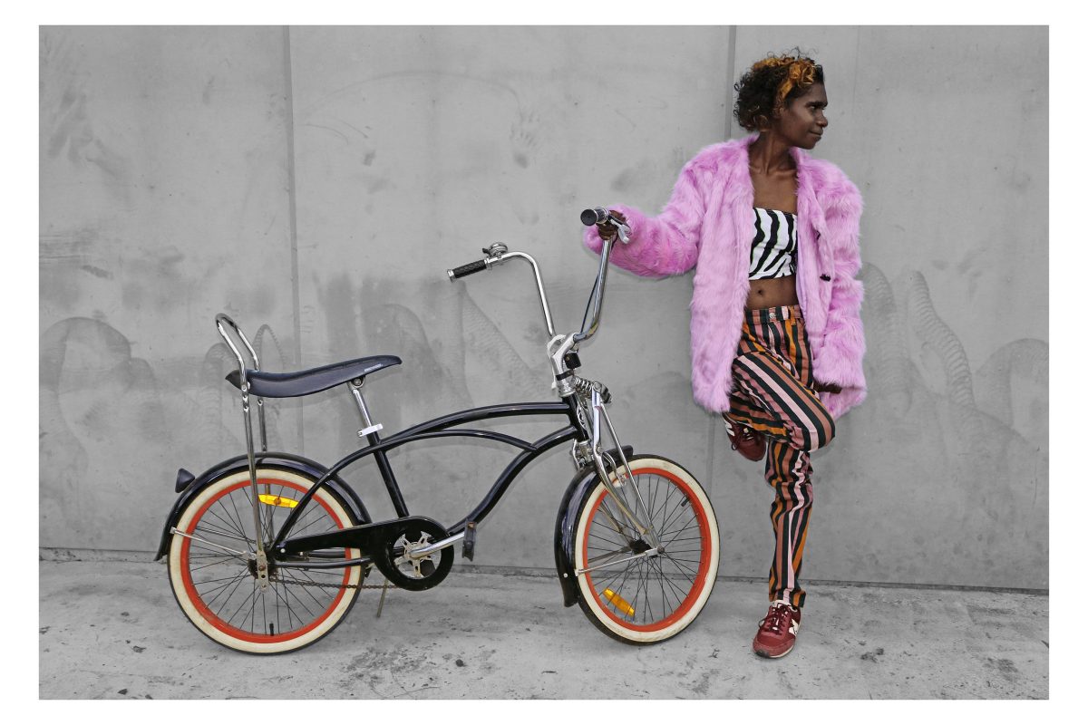 Girl wearing a pink coat holds onto a bike and leans against a wall. She and the bike are in colour and the rest of the image is in black and white.