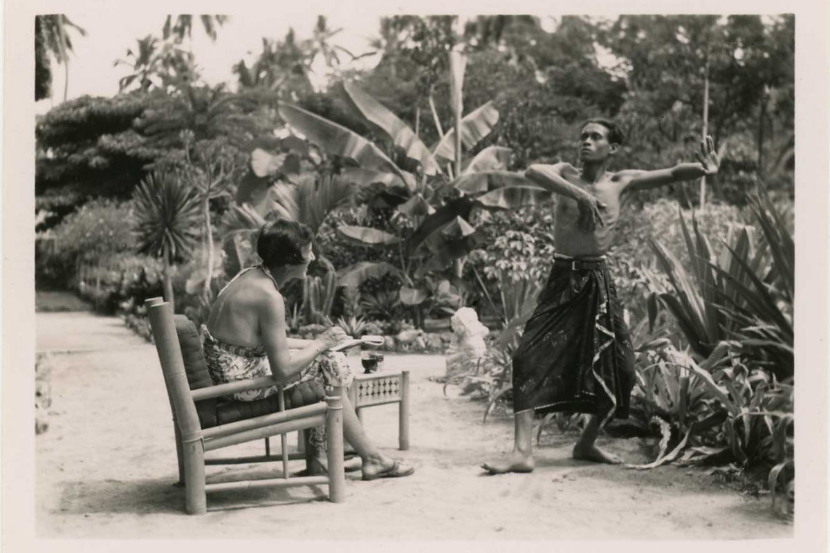 Archive photo of a woman sat on a chair watching as a man dances. The woman has a notebook and pen in her hands