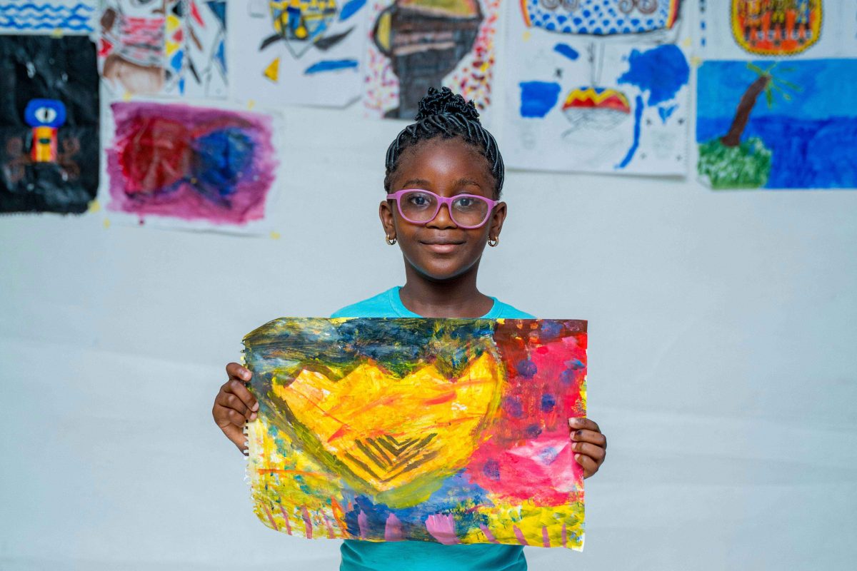 A child holds up a drawing in front of rows of children's artworks