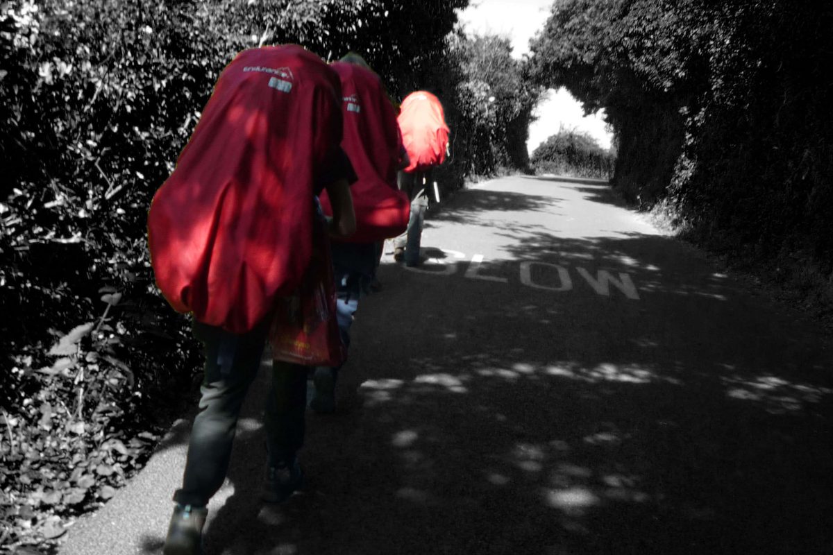 Children walk in a line down a country path, carrying large red backpacks. They are in colour and their surroundings are in black and white.