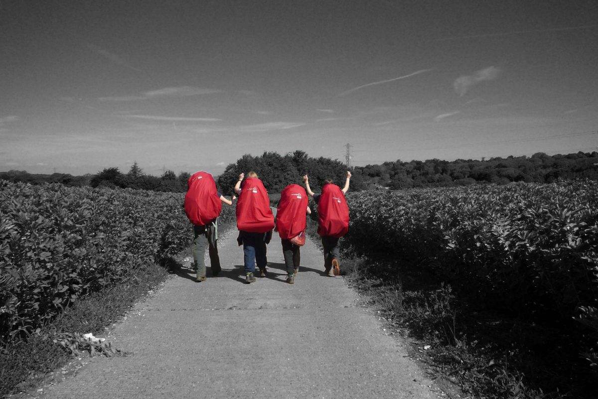 Four children stand in a line, carrying large red backpacks. They are in colour and their surroundings are in black and white.