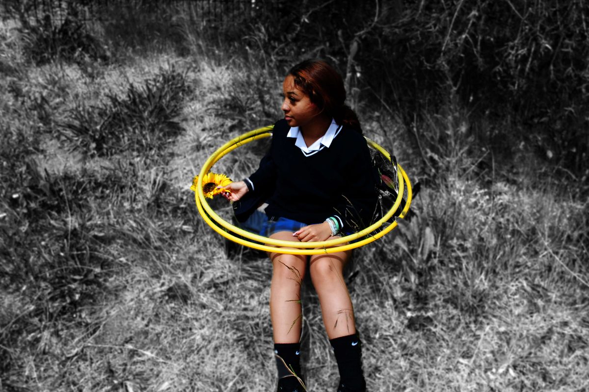 A girl sits on some grass with two yellow hula hoops placed around her. She is in colour and her surroundings are in black and white.