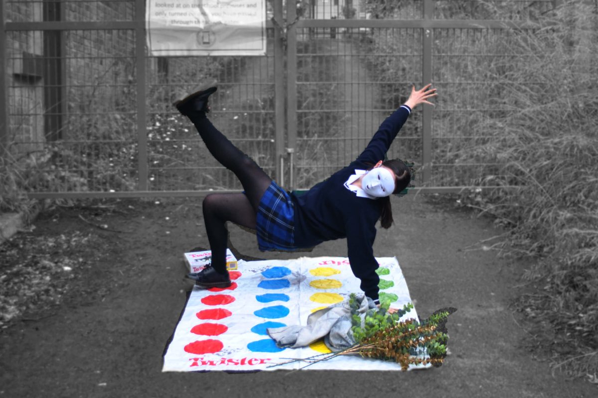 A girl wearing school uniform and a white mask plays on a Twister mat with one hand and one foot on the circles. There is a bunch of dried flowers next to her. She is in colour and her surroundings are in black and white.