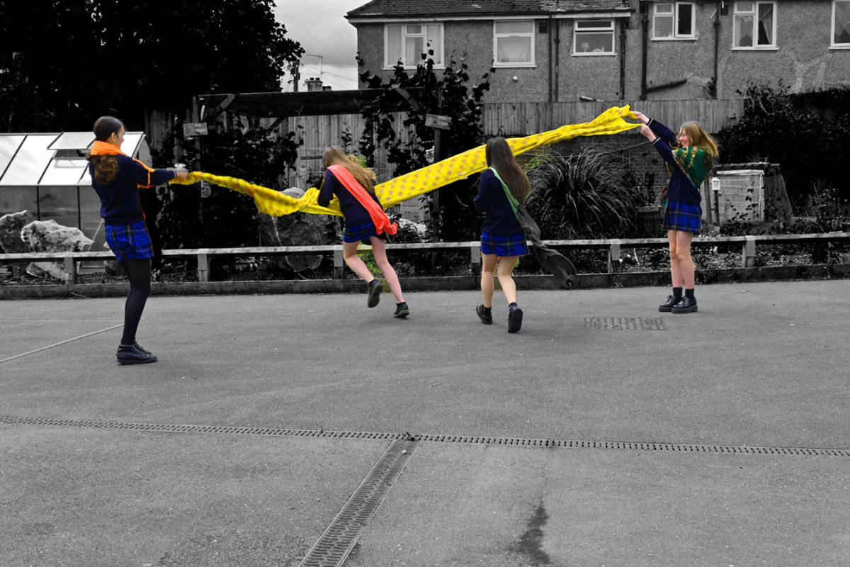 Four girls hold a piece of bright material in the air, with two in the middle running towards it. They are in colour and their surroundings are in black and white