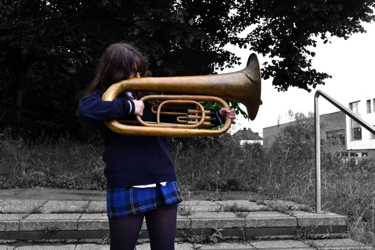 A child holds a tuba like a bow and arrow, pointing it off camera. She and the tuba are in colour and the surroundings are in black and white.